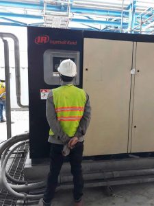 Kỹ thuật viên mặc áo phản quang đang kiểm tra thông số trên bảng điều khiển của máy nén khí trục vít Ingersoll Rand quy mô công nghiệp lớn.